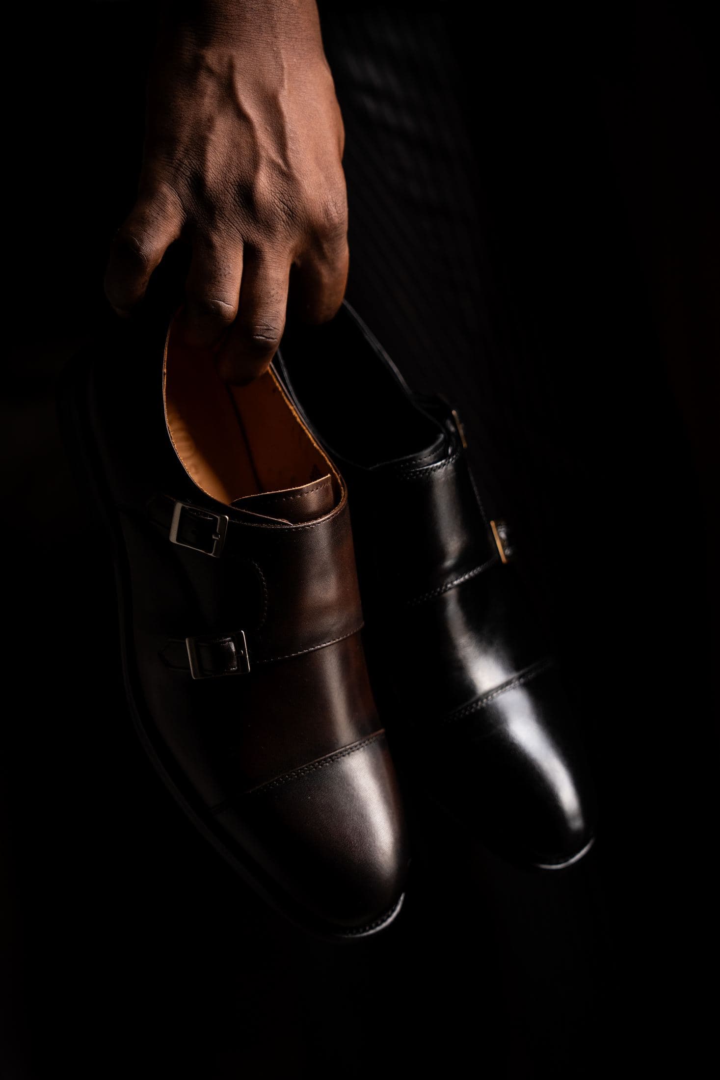 A hand lifting a pair of espresso double monk straps out of the dark